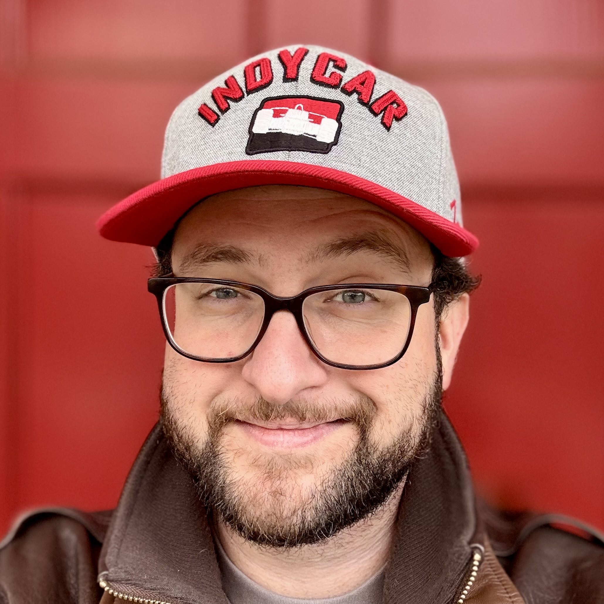 A man in a leather jacket, glasses, and an IndyCar hat